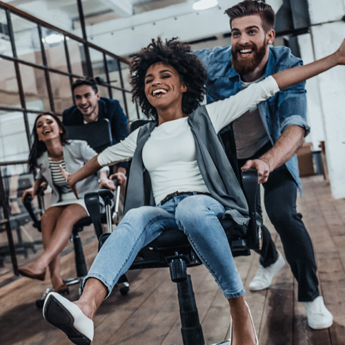 Coworkers smiling & racing in office rolling chairs, representing companies driving a positive brand
